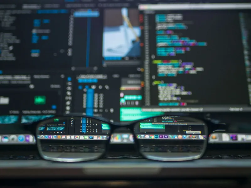 Black-framed eyeglasses resting on a laptop in front of blurred monitors showing source code and a video editing interface