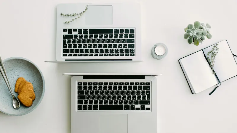 Two open silver laptops placed face to face on a white desk, seen from above, next to a notebook, a candle, a small succulent and a bowl of cookies