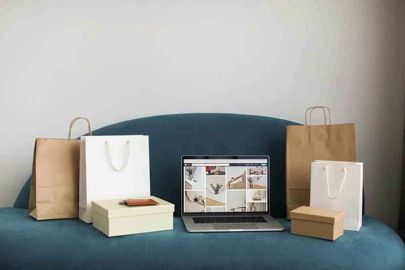 A laptop showing an online shop page on a teal sofa, surrounded by paper shopping bags and gift boxes