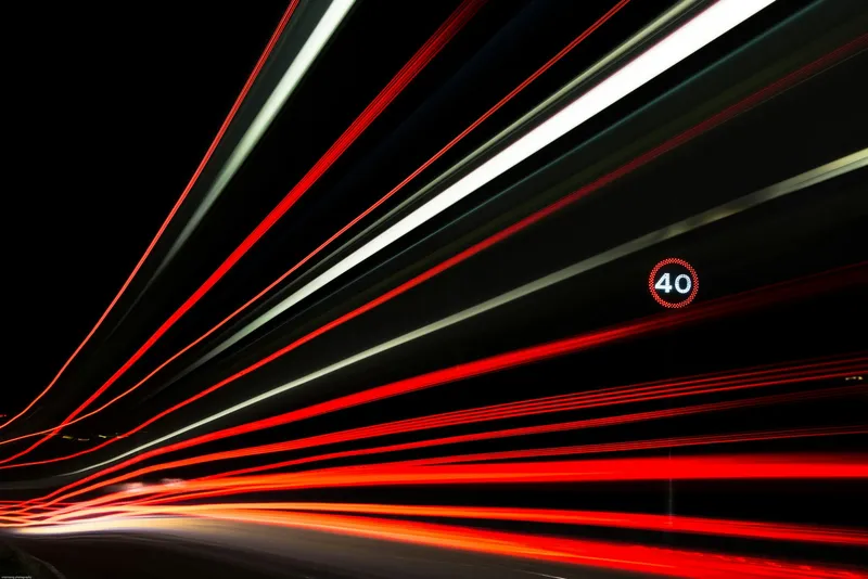 Long-exposure red and white light trails streaking past a 40 mph speed limit sign on a dark road at night